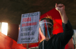 A demonstrator wearing a protective face shield and mask holds up a banner during a protest against Brazilian president, Jair Bolsonaro, in Sao Paulo July 26,. The banner reads “Out Bolsonaro, elections now!” Photo: CNS/Reuters