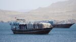 Two large wooden cargo boats anchored on calm blue sea with a rocky, arid shoreline in the distance.