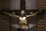 Statue of Jesus on the cross with a crown of thorns, arms outstretched, in a church setting with a stone wall behind.