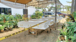 Outdoor funeral scene with three white caskets on wheeled stands under a canopy, surrounded by police tape and parked cars in the background.