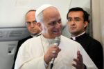 Pope Francis speaking into a handheld microphone, wearing white papal attire on a plane with two attendants in the background