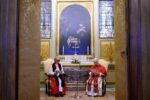 Two clergy members in liturgical robes sit and read from booklets in a richly decorated chapel with a large central painting behind them.