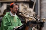 Catholic priest in a green vestment delivering a sermon at a lectern outdoors, speaking into a microphone.