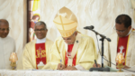 Catholic bishop in gold vestments signing a document at the altar during a ceremony, with two priests nearby and microphones present