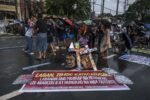 Crowd of protesters on a wet street, a large red banner with Filipino text on the ground, and a colorful cross sculpture atop two painted heads in the foreground.