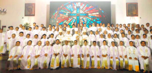 Group of priests in white liturgical robes posing for a portrait in a church, with a large stained-glass cross behind them. 80+ people in rows, front kneeling.