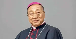Smiling elderly Catholic bishop wearing a pink zucchetto and black clerical robes with a red trim and glasses.