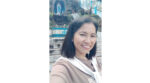 Smiling woman taking a selfie outdoors, wearing a brown shirt and light scarf, with stone steps and a blue railing behind her.