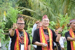 Group of people in traditional attire holding up leafy stems during an outdoor festival, with palm trees in the background.