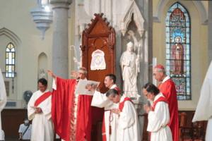 Group of Catholic priests in red and white liturgical vestments conducting a ceremony at the church front, with a carved lectern, statue, and stained-glass window behind them and a masked bystander in the background.