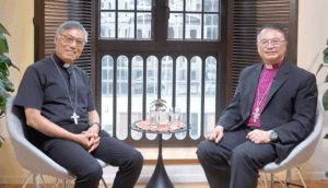 Two priests wearing clerical collars and cross necklaces sit in modern chairs on either side of a small round table with two glasses between them in a bright room.