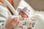 Newborn baby in a hospital bed being fed with a bottle, wrapped in a blanket with colorful footprints and wearing a striped bow hat.