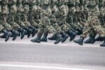 Soldiers in camouflage uniforms marching in formation on a paved street, black boots aligned in unison.