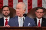 Elderly man in a blue pinstripe suit speaks at a podium with a microphone, flanked by two suited men, with a red-and-white striped backdrop.