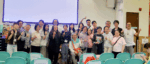 Group of about 25 people posing for a photo in a classroom with a projection screen, many smiling and giving thumbs-up gestures.