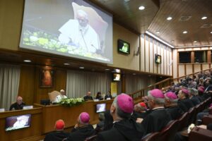 Panel of bishops and clergy seated at a long conference desk in a large meeting hall, with a projection screen showing a speaker in white robes and floral decor at the front.