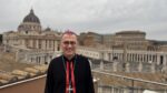 Catholic bishop in a black cassock with pink zucchetto and a cross necklace, standing on a rooftop terrace in Rome with St. Peter's Basilica in the background.