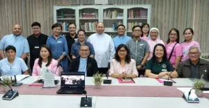 Group of 16 people posing for a photo around a conference table in a library-like room; front row seated with a laptop and documents, back row standing and smiling.