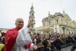 Catholic clergyman in red vestments waves to a crowd in front of a grand church with a tall bell tower.
