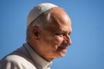 Close-up profile of an elderly man wearing a white kippah, beige jacket, against a bright blue sky.