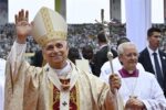 Catholic cleric wearing a gold-embroidered mitre waves to a crowd, holding a silver ceremonial staff in an outdoor ceremony with spectators in the background.