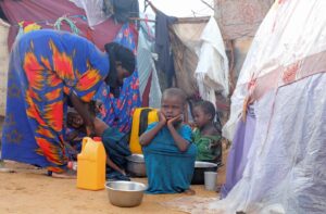 Young boy in a blue shirt sits on the dusty ground, hands on his cheeks, with bowls and a yellow jerrycan nearby in a makeshift camp.