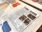 Front page of a vintage newspaper titled 'Sunday Examiner' on a glass display case with black-and-white photos and dense text blocks, surrounded by cards.