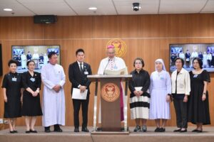 Ten people in formal attire stand on a stage behind a podium; a bishop in white robes and pink zucchetto reads from a binder, flanked by nine officials in front of a wood-paneled wall with emblems.