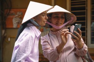 Two women in conical hats share a smartphone, looking at the screen together with masks hanging from their necks.
