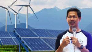 Man giving a talk with a microphone in front of solar panels and wind turbines in a rural renewable energy site. Memorandum-style presentation setting with mountains in the background.