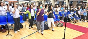 Large crowd standing and raising hands in a hall with blue chairs and a red carpet, suggesting a group activity or performance.