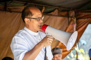 A clergyman wearing a white collar speaks into a red and white megaphone at an outdoor event.