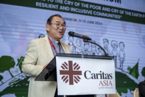 Man in a light beige suit speaks at a podium with a Caritas Asia sign and a microphone during a conference.