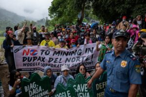 Crowd of protesters holding a banner that reads 'No to mining in Dupax del Norte; Woggle Corporation out now,' with a police officer in the foreground.