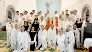 Group photo of Catholic clergy and nuns in white robes inside a church, posing for a formal gathering. Notable figures include a bishop in ornate vestments centered among priests and sisters.