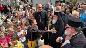 Orthodox priests greet a crowd of children and adults at an outdoor community gathering.