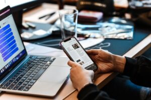 Person holds a smartphone displaying an article beside a laptop on a cluttered desk.