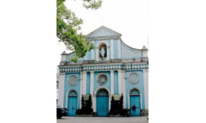 Blue-and-white church façade with three arched doors, circular windows, decorative columns, and a statue in a niche above the center door, partly shaded by a tree.