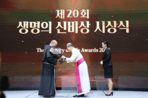 Two men on a stage at an awards ceremony exchange a bouquet; one wears black religious robes, the other white and pink clerical robes, with a suited assistant nearby.