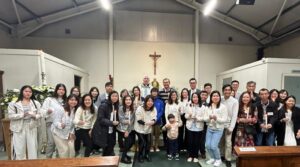 Group of people in a church hall posing for a photo around a crucifix altar, some holding lit candles, with a priest in the center.