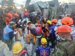 Rescue workers in helmets and uniforms search through debris at a collapsed building, coordinating amid onlookers nearby.