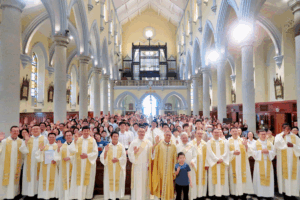 Group of priests in white robes with gold stoles posing at the altar, parishioners behind them waving inside a church hall with arches and an organ loft above.