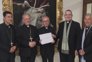 Five priests in black clerical suits stand together indoors; the central man holds a white envelope, with a gilded painting behind them.