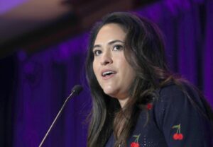 Woman speaking into a microphone on a stage with purple lighting, wearing a dark sweater with small cherry embroidery on the sleeve.