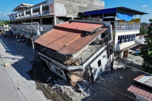 Aerial view of a multi-story building with a collapsed corner and rubble strewn across the street below in a urban area.