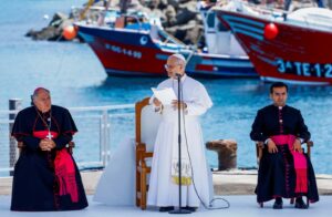 Pope speaking at a waterfront ceremony, holding papers at a mic, flanked by two bishops in black robes and pink sashes, boats in the harbor behind him.