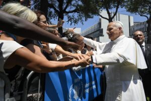 Pope Francis in white robes greets a crowd, shaking hands with supporters along a barrier outdoors, near a World Food Programme banner.