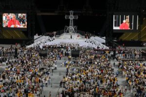 Large stadium ceremony with a crucifix centerpiece, white staircase, and an orchestra; thousands fill the arena.