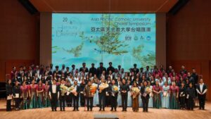 Large group of singers and presenters on a stage after a choral symposium, with a colorful backdrop and bouquets in hand.