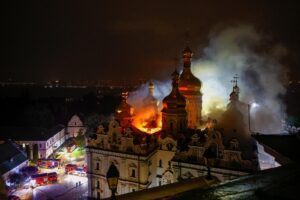 Nighttime cathedral with onion domes ablaze; flames and smoke rise from the roof while firetrucks line the street below.
